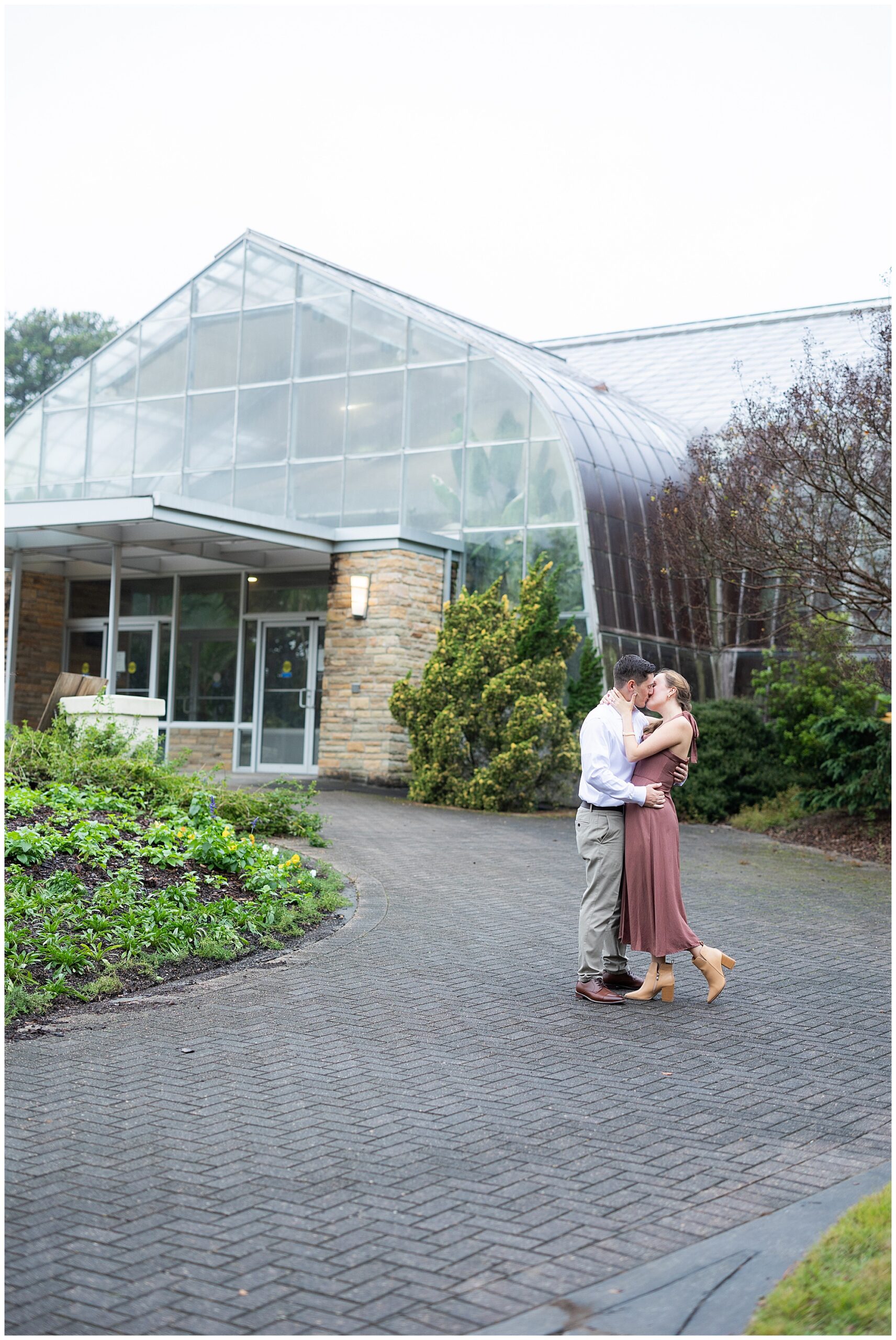 A black and white image of Catherine and Jake holding each other and kissing in front of the Botanical Garden's greenhouse. 