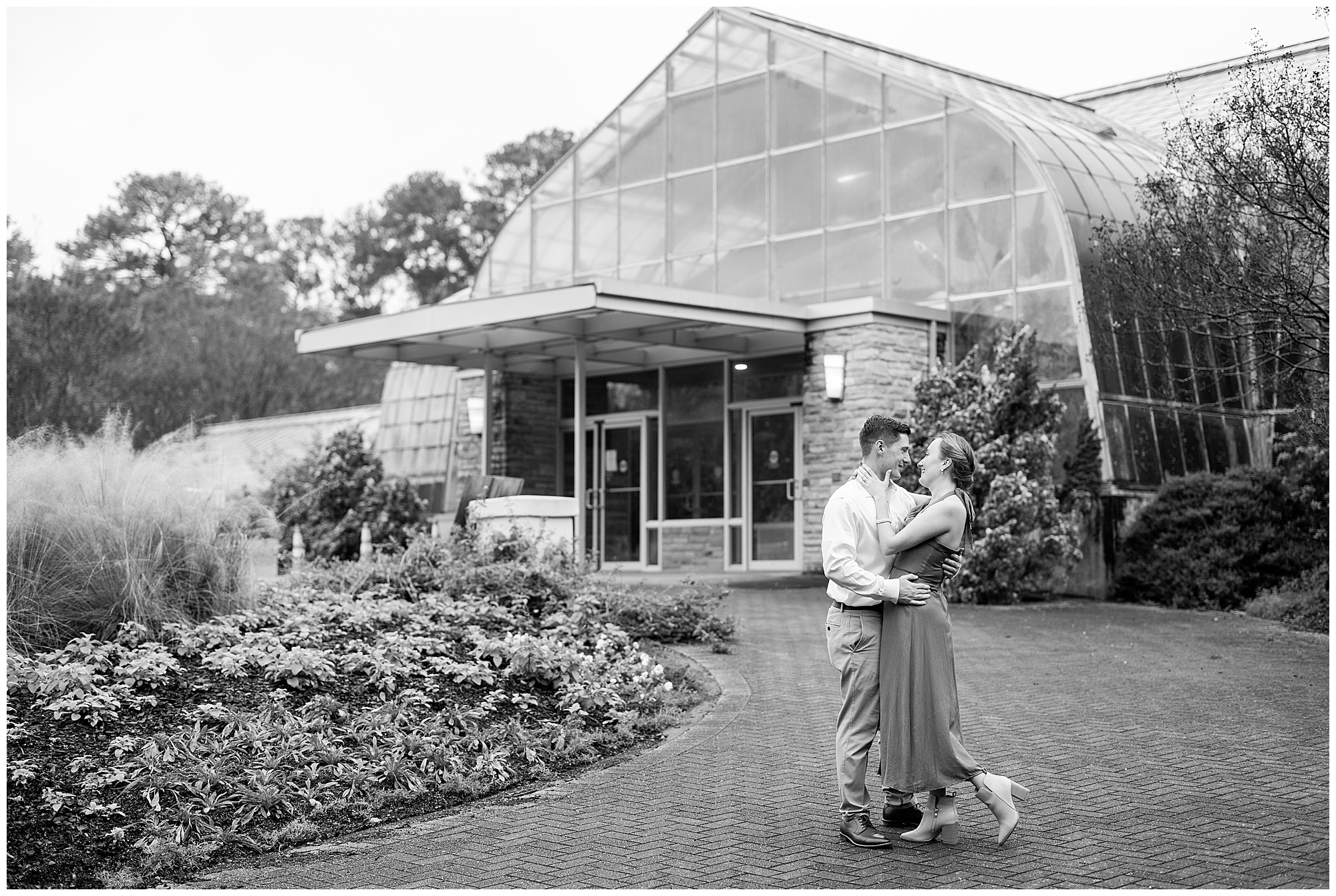 A black and white image of Catherine and Jake holding each other and smiling in front of the Botanical Garden's greenhouse. 
