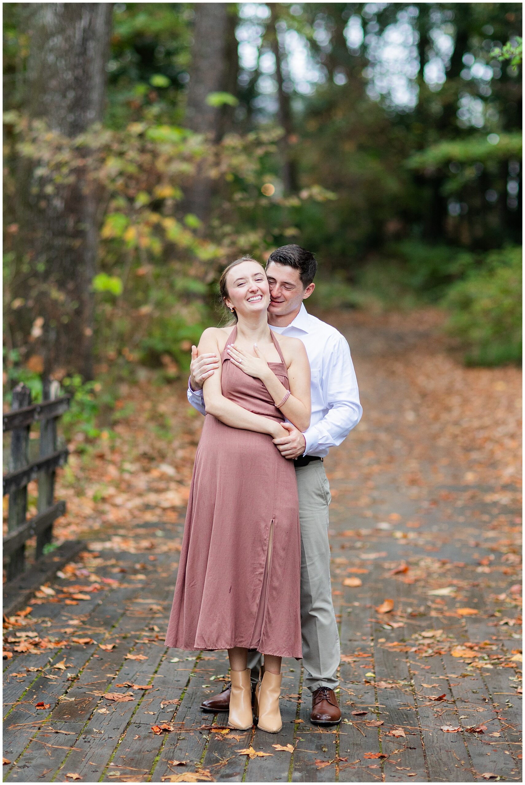 Jake holds Catherine from behind while they stand on a wooden bridge in the Botanical Gardens. They laugh together. 