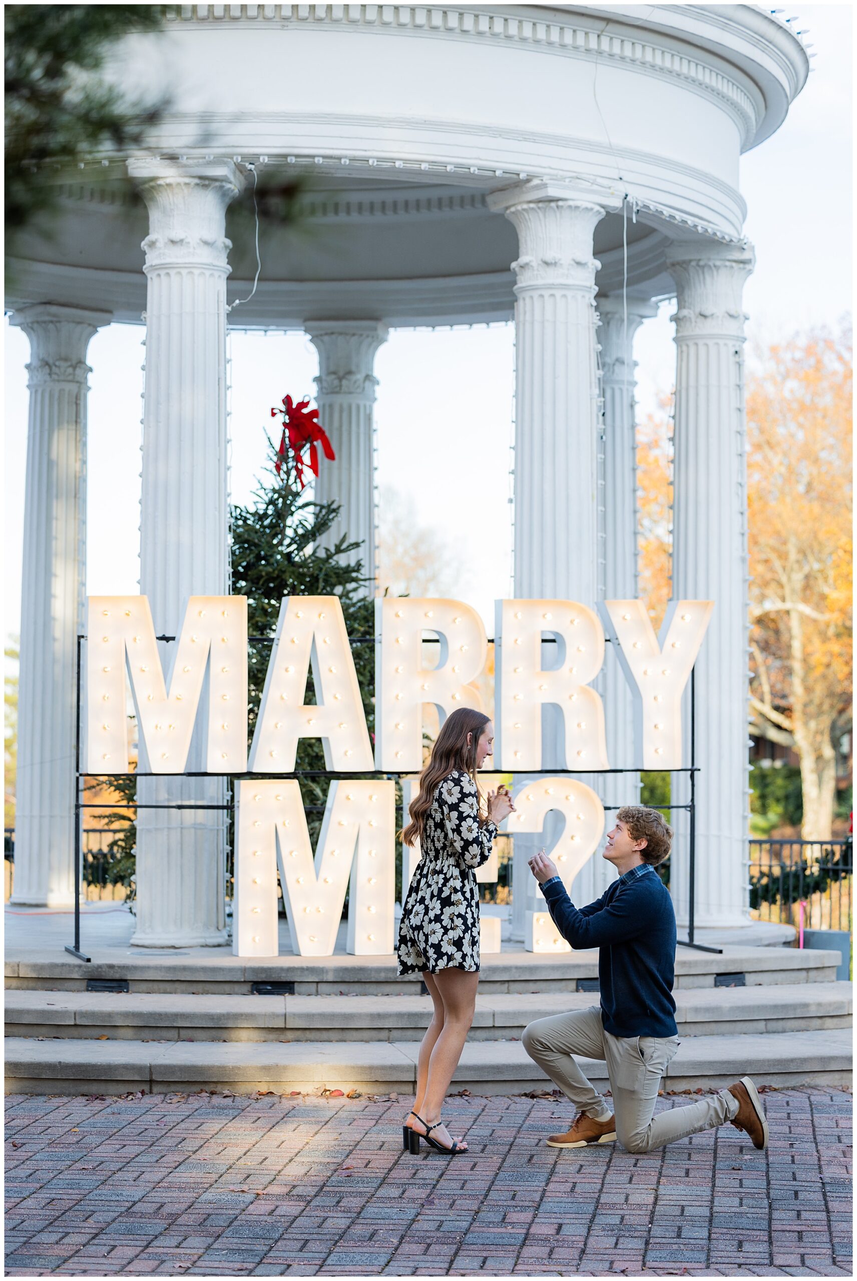 Camden is down on one knee, proposing to his girlfriend, Olivia. They stand in front of a large marquis sign that reads "Marry Me?" placed in from to Sibyl Temple.