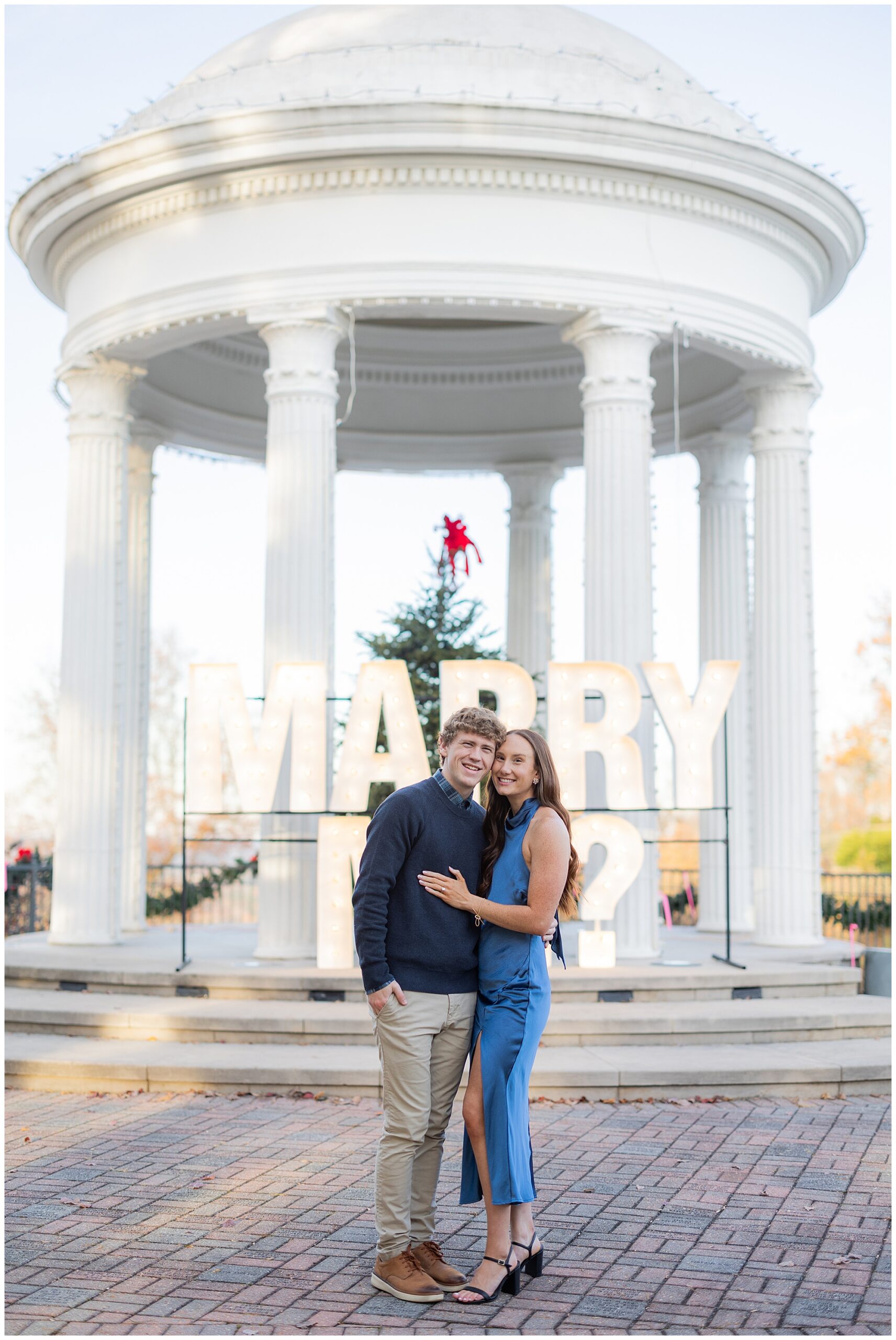 Camden and Olivia stand side by side and smile at the camera in front of the "Marry Me?" sign.